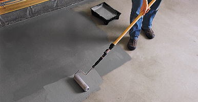 This image shows a man painting a garage floor with a roller brush.